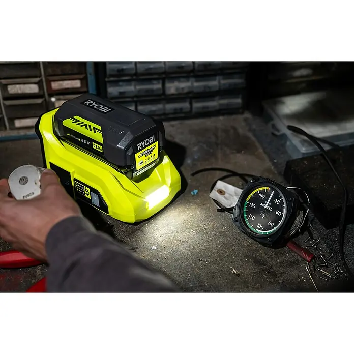 Ryobi Max Power Wechselrichter RY36BI300A-0 - 36 V, Ohne Akku, 300 W - Nahaufnahme eines RYOBI-Akkus mit hoher Kapazität, der eine Werkbank mit Werkzeugen und Messgeräten beleuchtet.