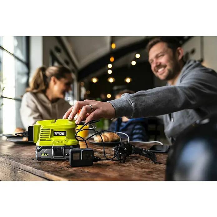 Ryobi ONE+ Wechselrichter RY18BI150A-0 - 18 V, Ohne Akku, 150 W - Nahaufnahme eines gelben Ryobi ONE+ Akku-Ladegeräts auf einem Holztisch in einem Café, lädt gleichzeitig eine GoPro und eine Smartwatch.