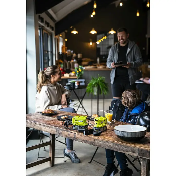 Ryobi ONE+ Wechselrichter RY18BI150A-0 - 18 V, Ohne Akku, 150 W - Ryobi-Akkus auf einem Holztisch in einem Café, umgeben von Menschen und Speisen. Die Akkus sind gelb und schwarz und mit Kabeln verbunden.