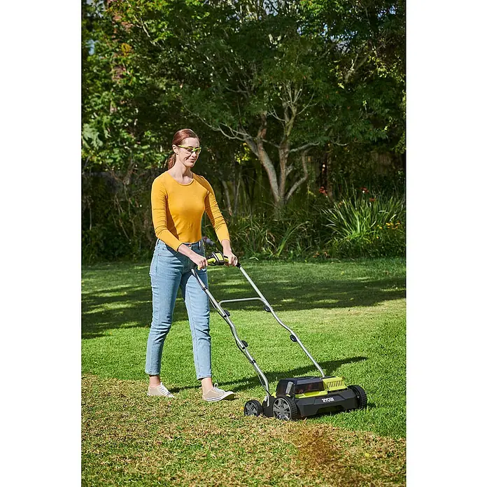 Ryobi ONE+ Escarificador de batería RY18SFX35A-0 - Número de baterías: Sin batería, 18 V, Anchura de trabajo: 35 cm - Mujer usa un aireador de césped Ryobi en el jardín. El aireador de césped es negro y amarillo y tiene un mango plateado.