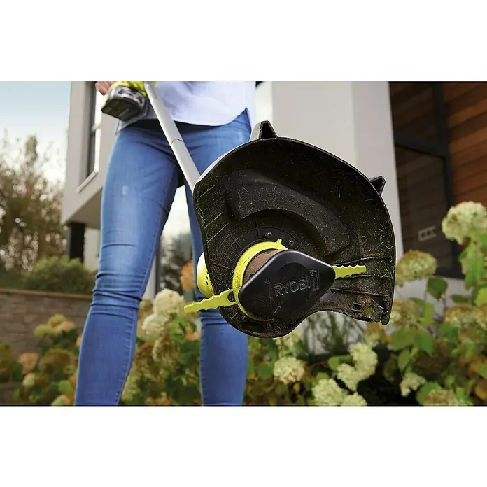 Ryobi Ersatzmesser RAC158 - Passend für: Ryobi-Zubehörteile RAC157 und RAC155 - Nahaufnahme eines Ryobi Rasentrimmers mit einem schwarzen Schutzschild und gelben Klingen vor dem Hintergrund einer Person und Vegetation.