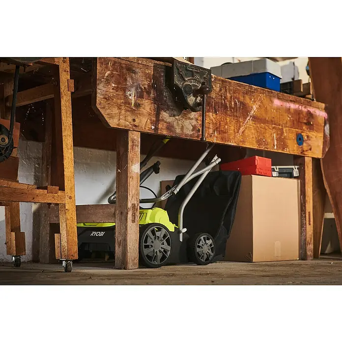 Ryobi ONE+ Akku-Vertikutierer RY18SFX35A-0 - Anzahl Akkus: Ohne Akku, 18 V, Arbeitsbreite: 35 cm - Eine Nahaufnahme eines RYOBI Rasenmähers, der unter einer hölzernen Werkbank mit diversen Werkzeugen und Kisten verstaut ist.