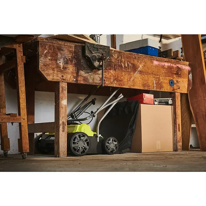 Ryobi ONE+ Akku-Vertikutierer RY18SFX35A-0 - Anzahl Akkus: Ohne Akku, 18 V, Arbeitsbreite: 35 cm - Eine Ryobi-Rasenmäher-Maschine steht unter einer Werkbank, zusammen mit einer Pappkiste und anderen Werkzeugen, in einer Garage.
