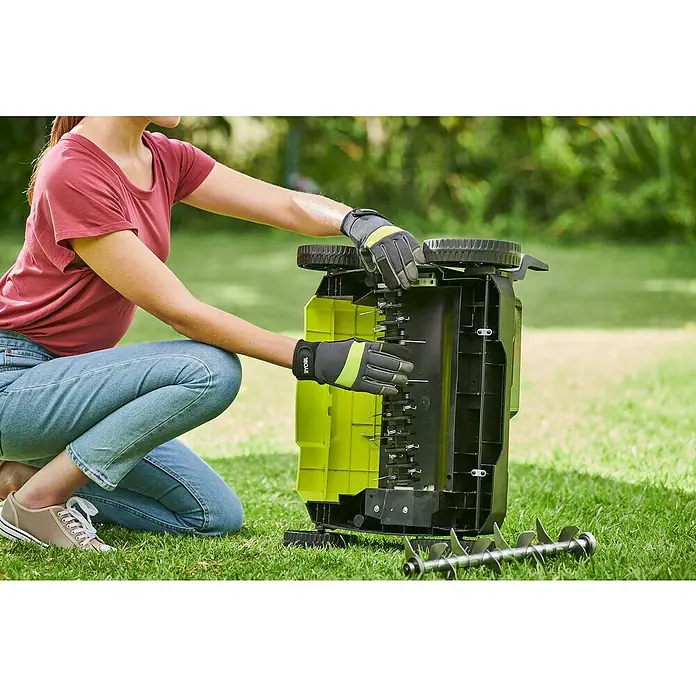 Ryobi ONE+ Escarificador de batería RY18SFX35A-0 - Número de baterías: Sin batería, 18 V, Anchura de trabajo: 35 cm - Una mujer inspecciona un escarificador Ryobi con guantes de trabajo negros y amarillos arrodillada en el césped.