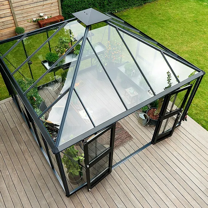 Juliana Gewächshaus Oase - Außenmaß inkl. Dachüberstand (B x T): 368 x 368 cm, Material Seitenwand: Glas, 3 mm, Anthrazit/Schwarz - Das Bild zeigt ein Gewächshaus mit Pflanzen, das auf einer Holzterrasse steht und von einem grünen Garten umgeben ist.