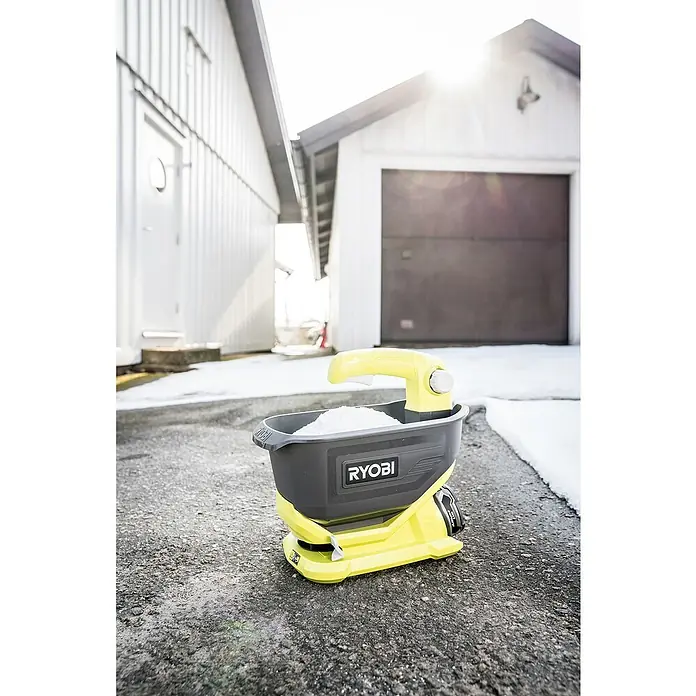 Ryobi ONE+ Handstreuer OSS1800 - 18 V, Anzahl Akkus: Ohne Akku - Nahaufnahme eines gelb-schwarzen Ryobi-Streuwagens mit Salz auf Asphalt vor Gebäuden, der sich ideal für die Eisbekämpfung eignet.