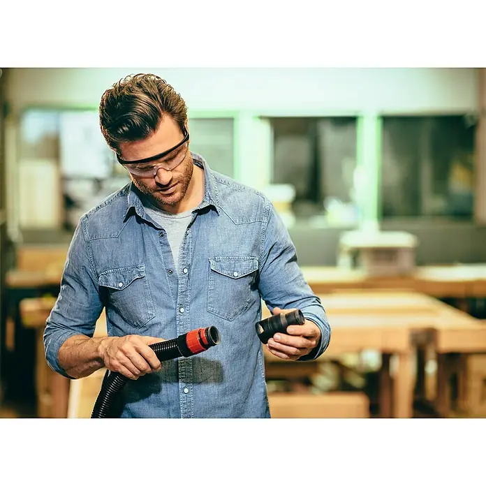 Bosch Elektro-Nass- & Trockensauger UniversalVac 15 - 1.000 W, Max. Unterdruck: 24.000 Pa, 15 l - Ein Mann mit Schutzbrille und Jeanshemd montiert einen Staubsaugerschlauch in einer Werkstatt. Werkzeuge sind im Hintergrund sichtbar.