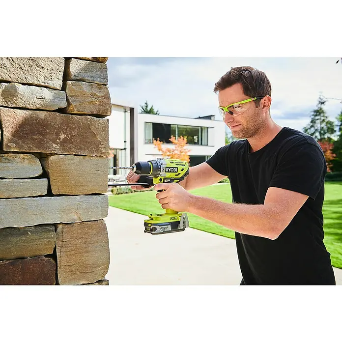 Ryobi ONE+ Akku-Schlagbohrschrauber R18PD7-252S - 18 V, 2 Akkus, 5 Ah, 85 Nm - Ein Mann bohrt mit einer RYOBI Bohrmaschine in eine Steinwand, Schutzbrille inklusive. Akkubetrieben für maximale Flexibilität.