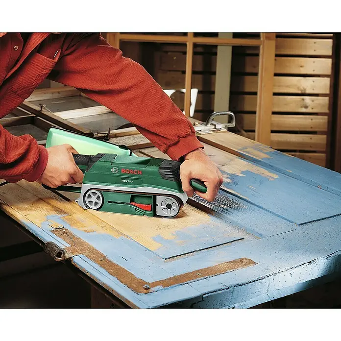 Bosch Elektro-Bandschleifmaschine PBS 75 A - 710 W, Bandgeschwindigkeit: 300 m/min - Nahaufnahme eines Mannes, der mit einem grünen Bosch-Bandschleifer eine Holztür abschleift.