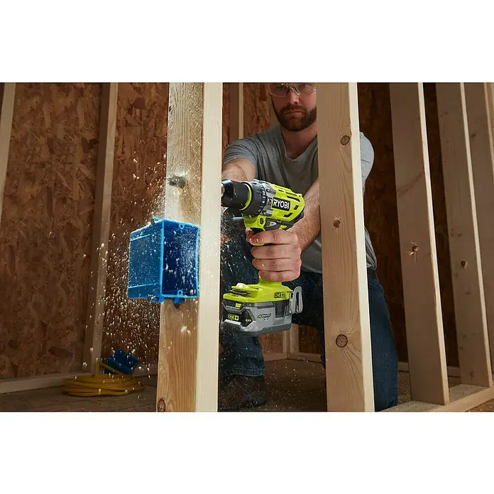 Ryobi ONE+ Akku-Schlagbohrschrauber R18PD7-252S - 18 V, 2 Akkus, 5 Ah, 85 Nm - Nahaufnahme eines Mannes, der mit einem gelb-schwarzen Ryobi-Akkuschrauber ein Loch in einen Holzrahmen bohrt.