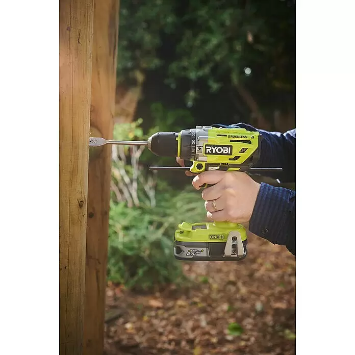Ryobi ONE+ Akku-Schlagbohrschrauber R18PD7-252S - 18 V, 2 Akkus, 5 Ah, 85 Nm - Eine Person bohrt mit einem gelb-schwarzen Ryobi Akku-Bohrschrauber und Flachfräsbohrer in einen Holzpfosten im Freien.