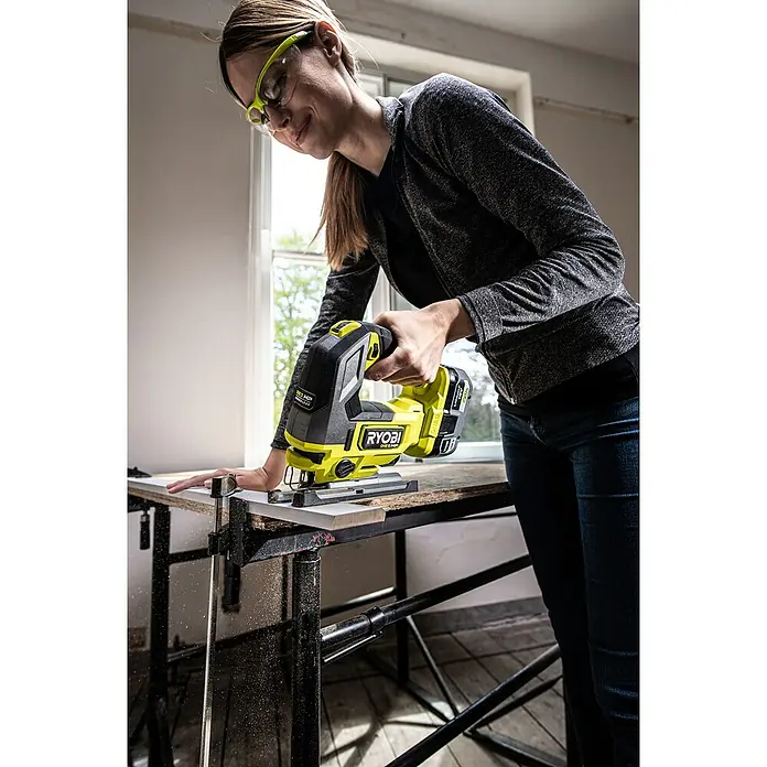 Ryobi ONE+HP Akku-Stichsäge RJS18X-0 Performance - 18 V, Ohne Akku, Leerlaufhubzahl: 800 /min - 3.400 /min - Frau benutzt eine RYOBI Stichsäge, um ein Holzstück zu sägen. Sie trägt eine Schutzbrille für die Arbeit.
