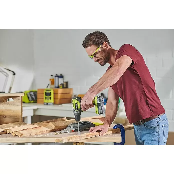 Ryobi ONE+ Akku-Schlagbohrschrauber R18PD5-0 - 18 V, Ohne Akku, 50 Nm - Mann mit Schutzbrille bohrt mit einem grünen Ryobi-Bohrer in Holz auf einer Werkbank.