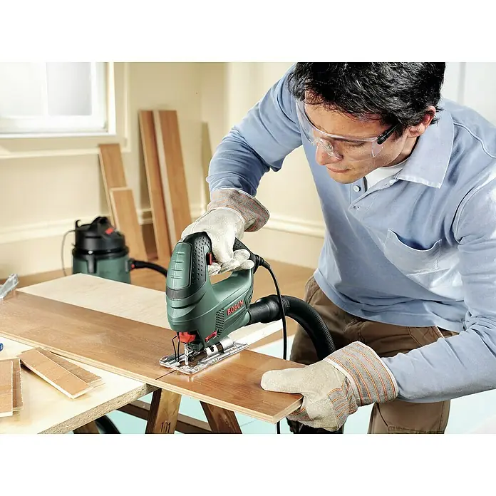 Bosch Elektro-Stichsäge PST 700 E - 500 /min - 3.100 /min, 70 mm, 500 W - Ein Mann benutzt eine Bosch Stichsäge, um ein Holzbrett zuzuschneiden, wobei er Schutzbrille und Handschuhe trägt. Staubabsaugung ist auch vorhanden.
