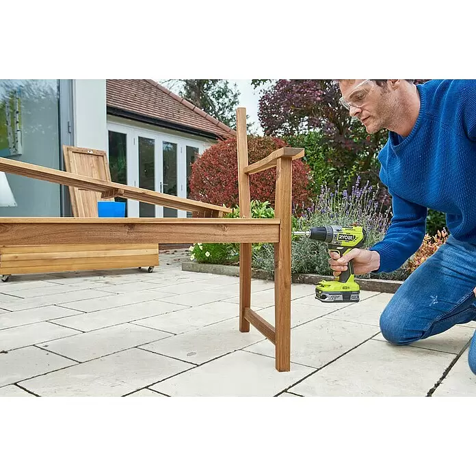 Ryobi ONE+ Akku-Schlagbohrschrauber R18PD5-0 - 18 V, Ohne Akku, 50 Nm - Person montiert eine braune Holz-Gartenliege mit einem Bohrer auf einer Terrasse.