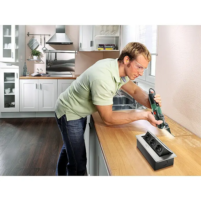 Bosch Elektro-Multifunktionswerkzeug PMF 350 CES - 350 W, STARLOCK-System - Ein Mann benutzt ein Multifunktionswerkzeug, um eine Steckdose in eine Küchenarbeitsplatte einzubauen. Das Werkzeug schneidet präzise in das Holz.