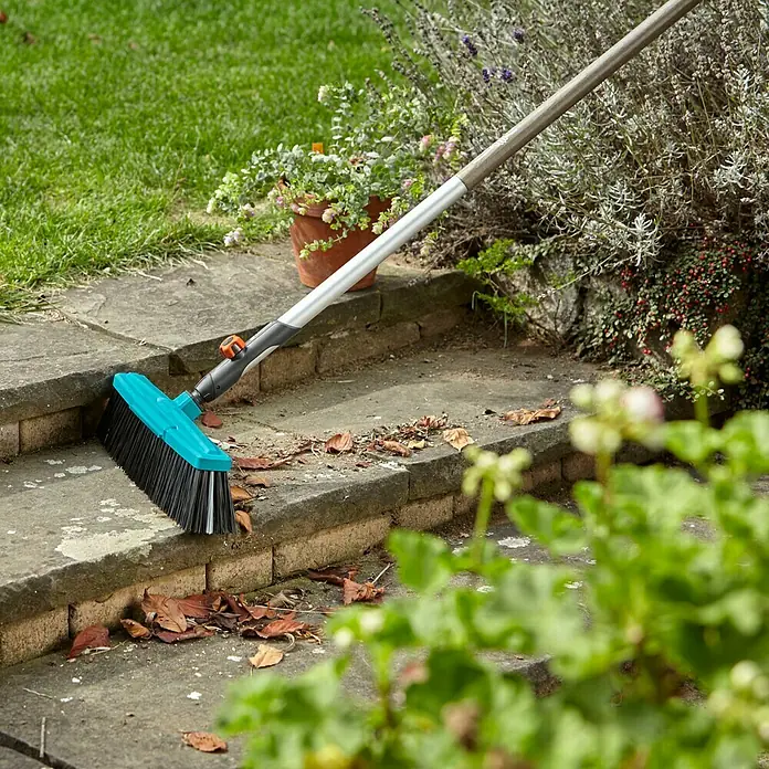 Gardena Combisystem Besen - Arbeitsbreite: 45 cm, Material Borsten: Polypropylen - Eine blaue Kehrbürste mit Teleskopstiel säubert eine Steintreppe von Laub. Der Garten im Hintergrund ist begrünt mit Sträuchern und Blumen.