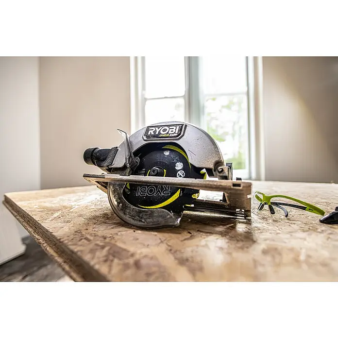 Ryobi ONE+HP Sierra circular de batería RCS18X - 18 V, Sin batería, 4.500 r.p.m. - Primer plano de una sierra circular Ryobi ONE+ sobre una superficie de madera, con gafas de seguridad al lado, sobre un fondo luminoso.