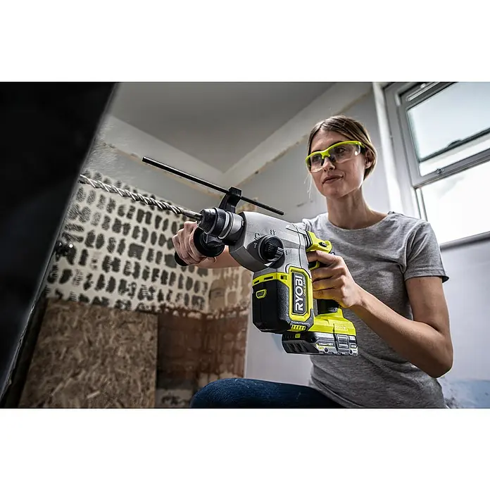 Ryobi ONE+HP Akku-Kombihammer RSDS18X-0 - 18 V, Ohne Akku, 2,5 J - Nahaufnahme einer Frau, die eine Ryobi Akku-Bohrmaschine benutzt, mit Schutzbrille und Staub in der Luft, während sie durch eine Wand bohrt.
