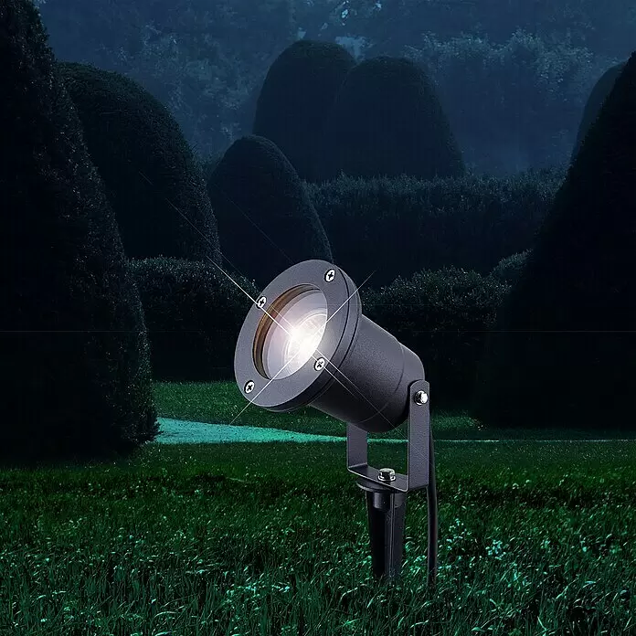 LED-Gartenstrahler Splash - 3 W, Grau, IP44, LED - Schwarzer Außenstrahler mit beleuchteter Glühbirne auf einem Erdspieß, der eine dunkle Gartenlandschaft beleuchtet.