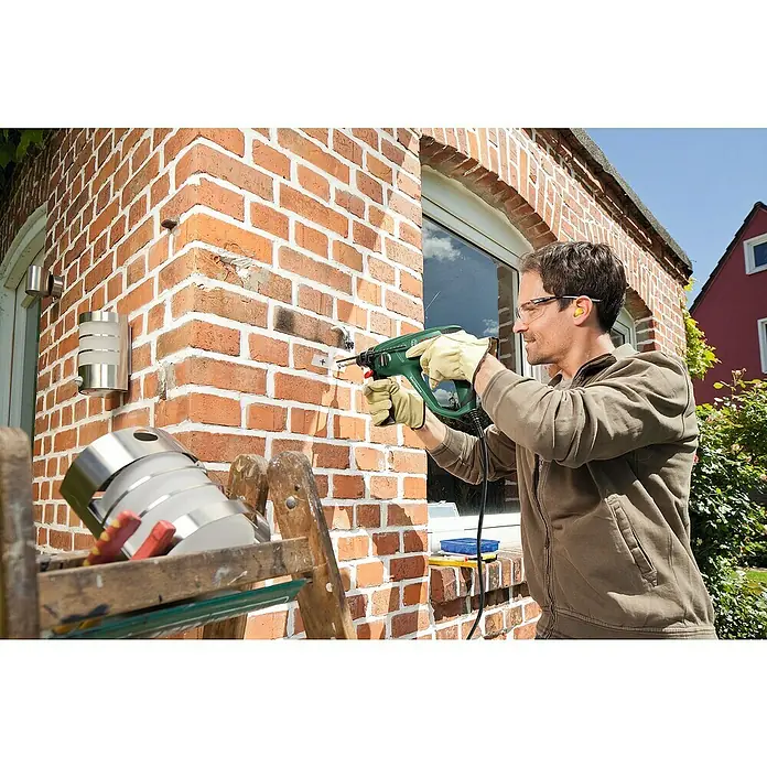 Bosch Kombihammer PBH 2200 RE - 570 W, 1,8 J, SDS-Plus-Aufnahme - Ein Mann bohrt mit einer grünen Bohrmaschine in eine Ziegelwand. Er trägt Schutzbrille und Handschuhe für die Arbeitssicherheit.