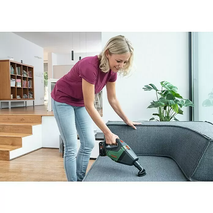 Bosch Power for All 18V Akku-Handstaubsauger UniversalVac 18 - 18 V, 1 Akku, 2,5 Ah, Behältervolumen: 0,5 l - Frau reinigt ein graues Sofa mit einem dunkelgrünen und schwarzen Bosch Handstaubsauger.