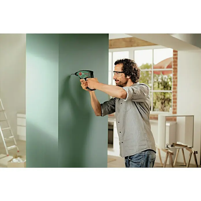 Bosch Power for All 12V Akku-Bohrhammer Uneo - 12 V, 1 Akku, 2 Ah, 0,5 J - Ein Mann bohrt mit einer grünen Bosch-Bohrmaschine ein Loch in eine mintgrüne Wand, während er eine Schutzbrille trägt.