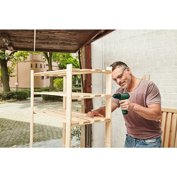 Bosch Power for All 12V Akku-Bohrschrauber EasyDrill 1200 - 12 V, 1 Akku, 1,5 Ah, 30 Nm - Ein Mann baut ein hölzernes Regal mit einem grünen Bosch-Akkuschrauber. Das Regal steht draußen vor einer Ziegelwand und Bäumen.
