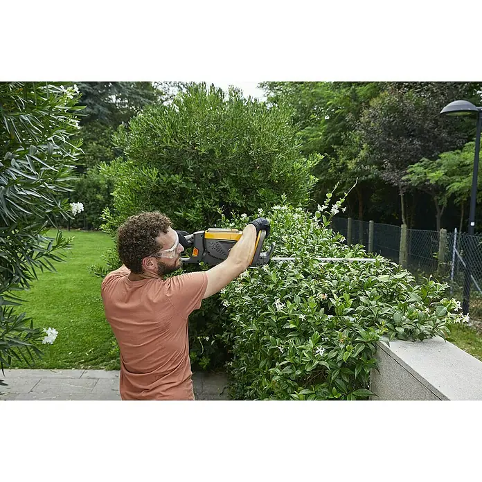 Stiga 500er Serie Akku-Heckenschere HT 500e Set48 V, Li-Ionen, 1 Akku, Schnittlänge: 58 cm - Ein Mann schneidet mit einer gelben Heckenschere eine Hecke. Er trägt Schutzbrille und Handschuhe.