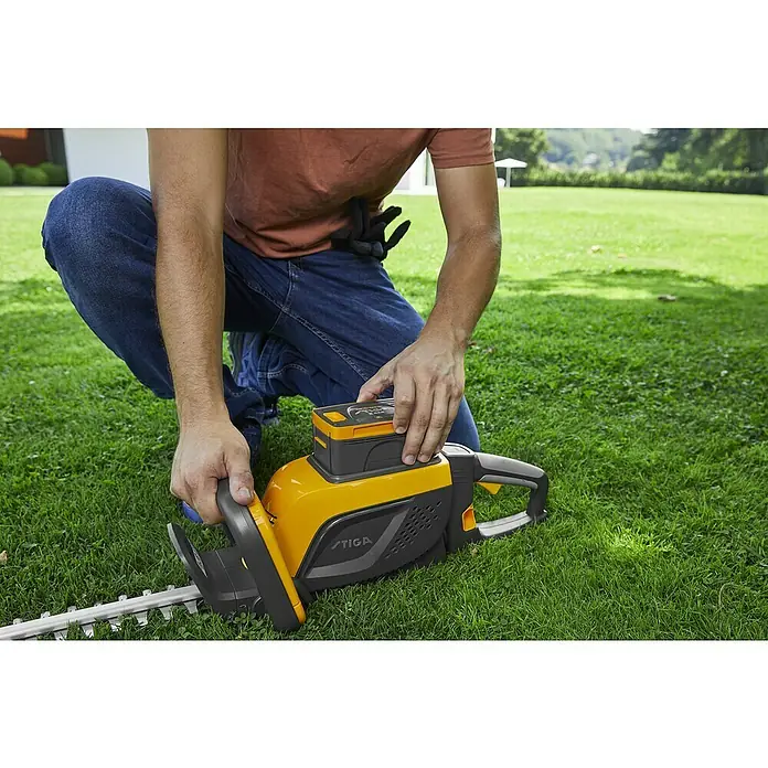 Stiga 500er Serie Akku-Heckenschere HT 500e Set - 48 V, Li-Ionen, 1 Akku, Schnittlänge: 58 cm - Ein Mann kniet im Gras und bedient eine gelbe STIGA-Heckenschere. Das Gerät ist mit einem Akku ausgestattet.