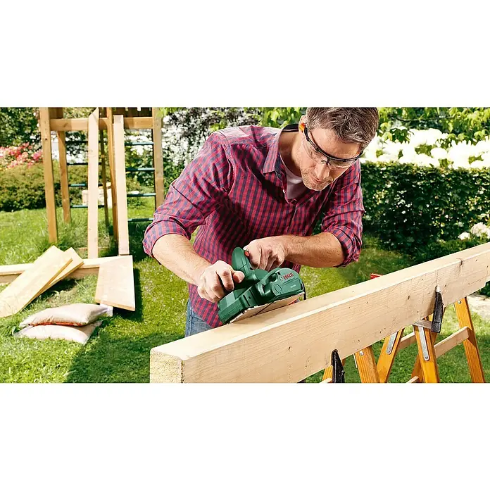 Bosch Handhobel PHO 1500 - 550 W, Hobeltiefe: 0 mm - 1,5 mm, 19.500 U/min - Ein Mann benutzt einen Bosch Elektrohobel, um ein Holzbrett im Freien zu bearbeiten. Er trägt eine Schutzbrille für die Arbeitssicherheit.