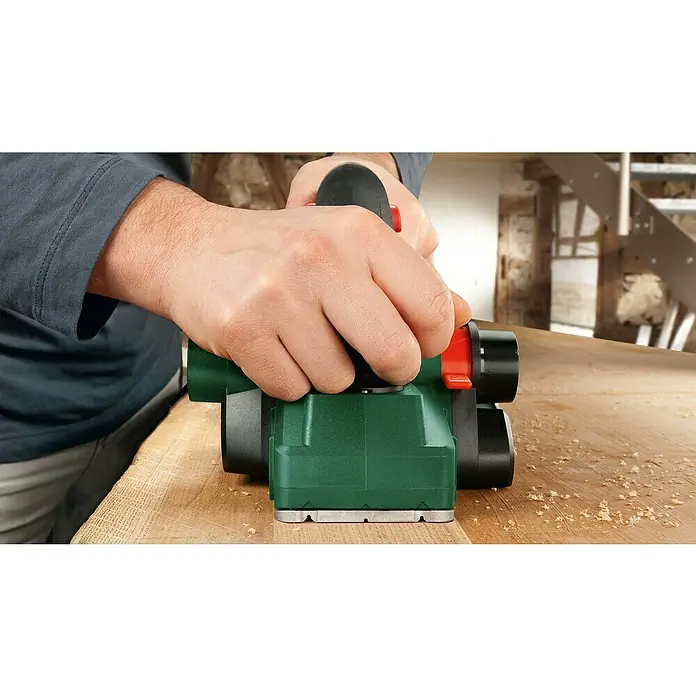 Bosch Elektrohobel PHO 2000 - 680 W, Hobeltiefe: 0 mm - 2 mm - Nahaufnahme eines Hobels in Aktion, der Holzspäne erzeugt, während er über ein Holzbrett gleitet, um eine glatte Oberfläche zu schaffen.