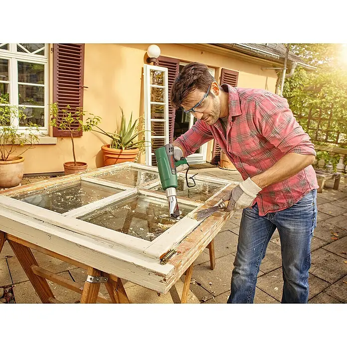 Bosch Heißluftgebläse EasyHeat 500 - 1.600 W, Temperaturbereich: 300 °C - 500 °C - Mann entfernt mit einem elektrischen Farbschaber Farbe von einem Fensterrahmen, um ihn für die weitere Bearbeitung vorzubereiten.