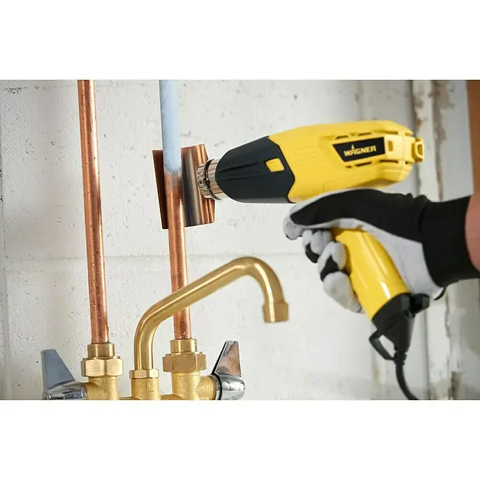 Wagner Heißluftgebläse Furno 300 - Leistungsaufnahme: 1.600 W, Temperaturbereich: 400 °C - 575 °C - Nahaufnahme einer Hand, die ein gelbes Wagner-Heißluftgebläse zur Bearbeitung von Kupferrohren verwendet. Das Werkzeug erhitzt ein Lotstück.