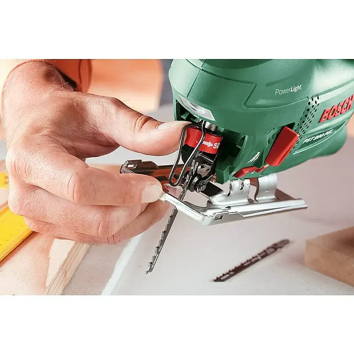 Bosch Elektro-Pendelhubstichsäge PST 900 PEL - 500 /min - 3.100 /min, Max. Schnitttiefe in Holz: 90 mm, Anzahl Pendelhubstufen: 4 Stk. - Ein Mann wechselt das Sägeblatt an einer grünen Bosch PST 900 PEL Stichsäge mit rotem Schnellspannhebel. Das Werkzeug ist bereit für präzise Holzarbeiten.