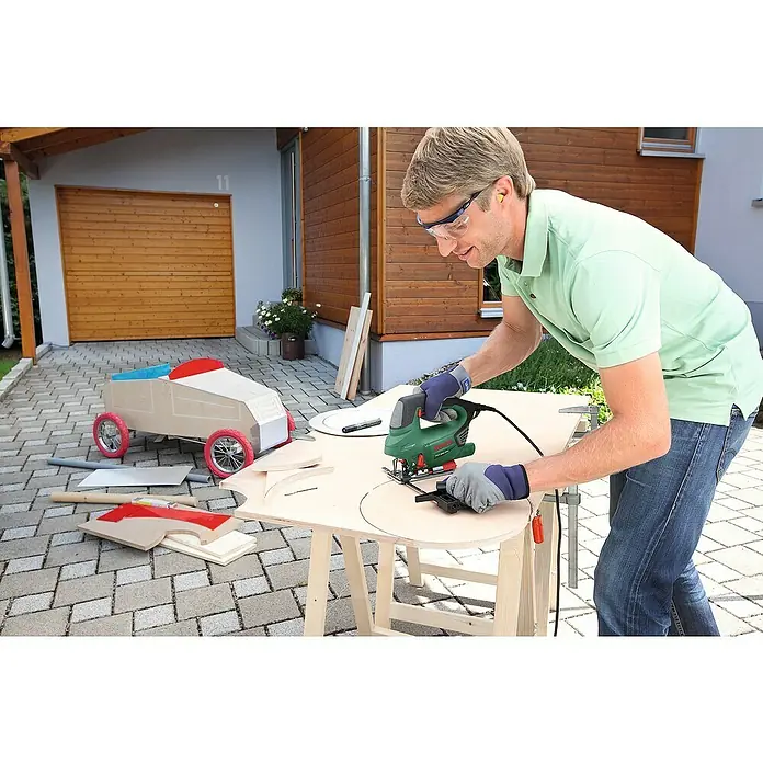 Bosch Elektro-Pendelhubstichsäge PST 900 PEL - 500 /min - 3.100 /min, Max. Schnitttiefe in Holz: 90 mm, Anzahl Pendelhubstufen: 4 Stk. - Ein Mann sägt mit einer Stichsäge ein Holzbrett, um ein Spielzeugauto zu bauen. Er trägt dabei Sicherheitsausrüstung.