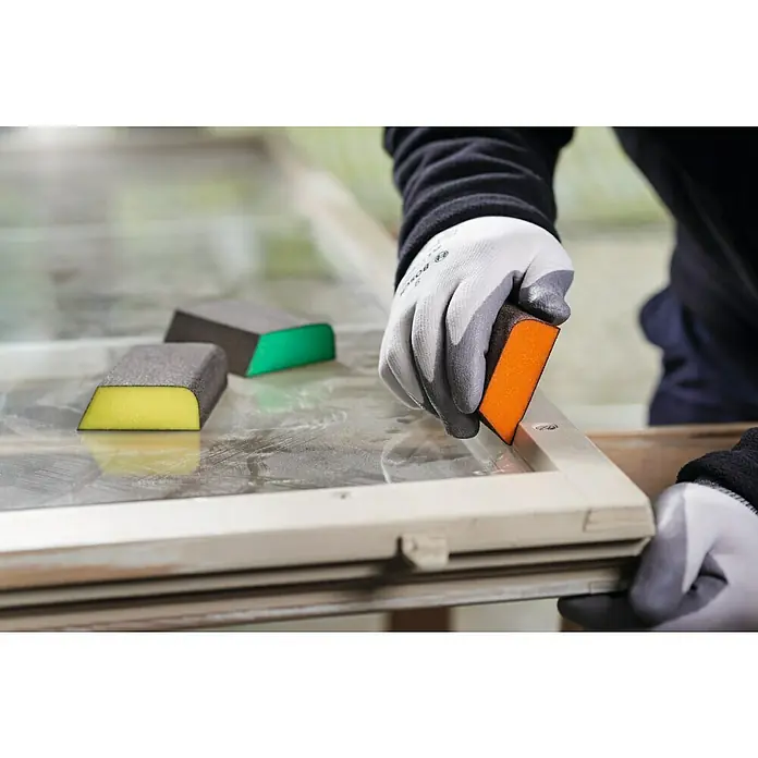 Bosch Professional Expert Schleifschwamm S470 - Korngröße: Mittel, L x B x H: 97 x 69 x 26 mm, 3 -tlg. - Eine Person schleift einen Fensterrahmen mit einem Schleifklotz, während andere Schleifklötze in Gelb und Grün daneben liegen.
