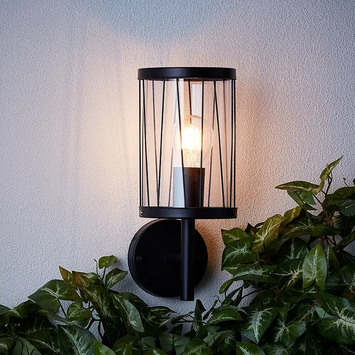 Brilliant Reed Außenwandleuchte - 60 W, Ø x H: 13,3 x 31,2 cm, Schwarz matt, E27 - Elegante Wandleuchte mit schwarzem Metallgestell und klarem Glaszylinder, umgeben von üppigem Grün, sorgt für stimmungsvolle Beleuchtung.