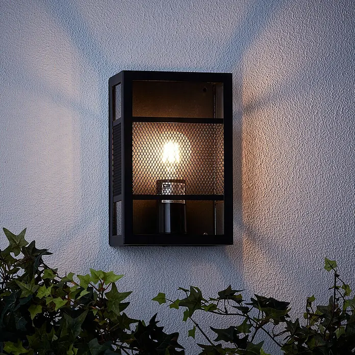 Brilliant Getta Außenwandleuchte - 20 W, 10 x 17 x 26 cm, Schwarz, IP44 - Eine elegante schwarze Wandleuchte mit einem Gitterdesign, die warmes Licht an eine strukturierte Wand wirft und von Grün umrahmt wird.