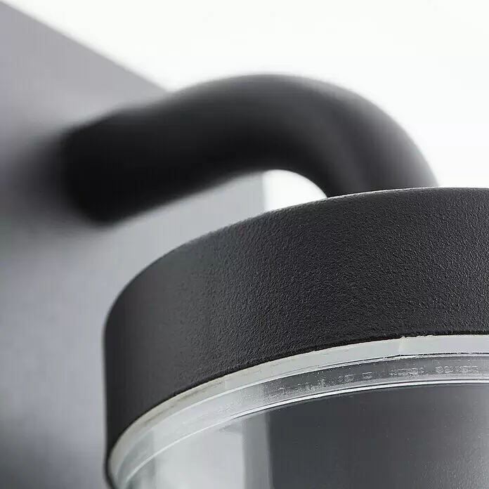 Brilliant Außenwandleuchte Aosta - 60 W, 12 x 8,5 x 30,6 cm, Schwarz matt, IP44, Hängend - Nahaufnahme einer schwarzen, texturierten Außenleuchte oder eines Sensors mit klarem, transparentem Unterteil.