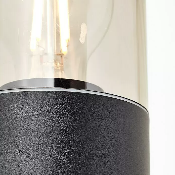 Brilliant Sensor-Außenleuchte Sergioro - 60 W, 12,5 x 9 x 33,2 cm, Schwarz matt, IP44 - Nahaufnahme einer modernen Lampe mit strukturierter dunkelgrauer Basis, silbernem Ring und klarem Glaszylinder mit leuchtender Glühbirne.