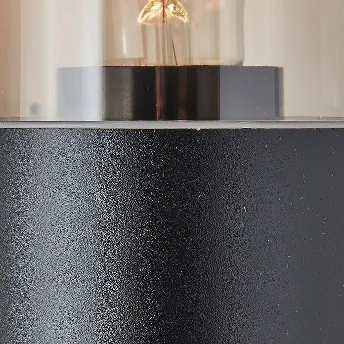 Brilliant Außenwandleuchte Sergioro - 18 W, 12,5 x 9 x 33,2 cm, Schwarz matt, IP44 - Nahaufnahme einer eleganten schwarzen Tischleuchte mit einer durchsichtigen Glühbirne, die ein warmes Licht ausstrahlt.