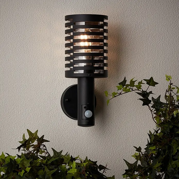 Brilliant Venlo Sensor-Außenleuchte - 18 W, Ø x H: 12,7 x 32,7 cm, Schwarz matt, IP44 - Eine moderne schwarze Außenwandleuchte mit Bewegungsmelder, die an einer Wand mit Efeu befestigt ist.