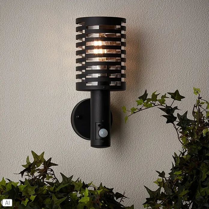 Brilliant Venlo Sensor-Außenleuchte - 18 W, Ø x H: 12,7 x 32,7 cm, Schwarz matt, IP44 - Eine moderne schwarze Außenwandleuchte mit Bewegungsmelder, die an einer Wand mit Efeu befestigt ist.
