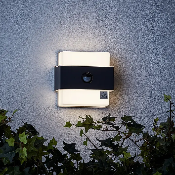 Brilliant LED-Sensor-Außenwandleuchte Archie - 7,5 W, L x B x H: 7 x 14,5 x 15 cm, Anthrazit, Neutralweiß - Eine quadratische Außenleuchte mit Bewegungsmelder an einer Wand, umgeben von Pflanzen. Sie ist weiß und schwarz.