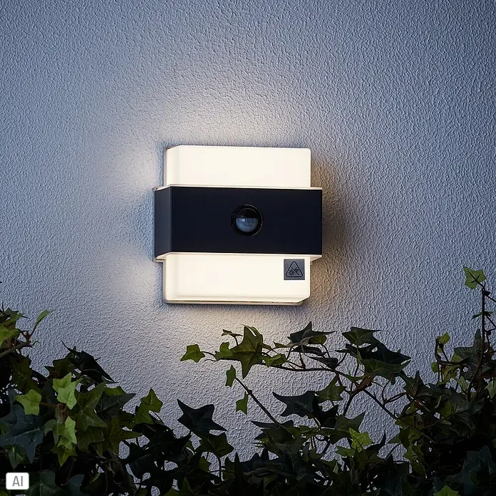 Brilliant LED-Sensor-Außenwandleuchte Archie - 7,5 W, L x B x H: 7 x 14,5 x 15 cm, Anthrazit, Neutralweiß - Eine quadratische Außenleuchte mit Bewegungsmelder an einer Wand, umgeben von Pflanzen. Sie ist weiß und schwarz.