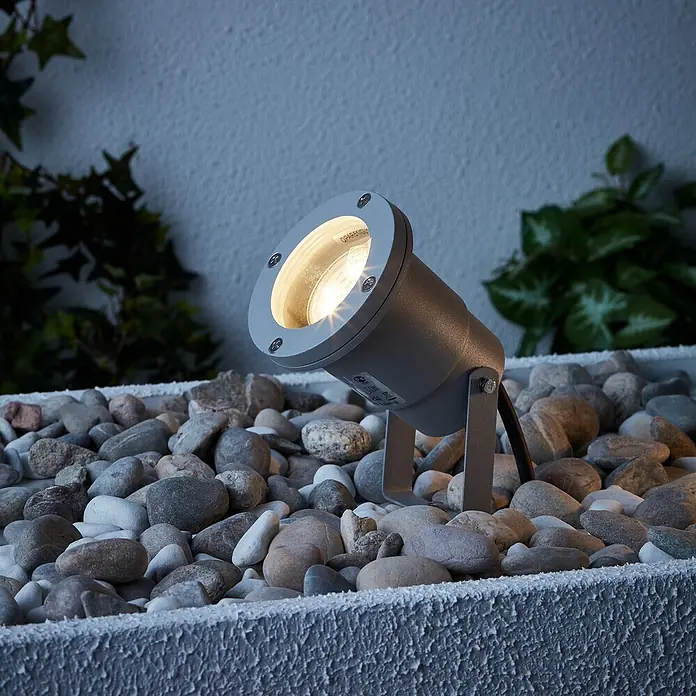 Brilliant LED-Erdspießleuchte Janko - 3 W, Ø x H: 9 x 16 cm, Grau, IP44 - Eine graue Gartenleuchte auf Kies mit warmweißem Licht für eine stimmungsvolle Beleuchtung im Außenbereich.