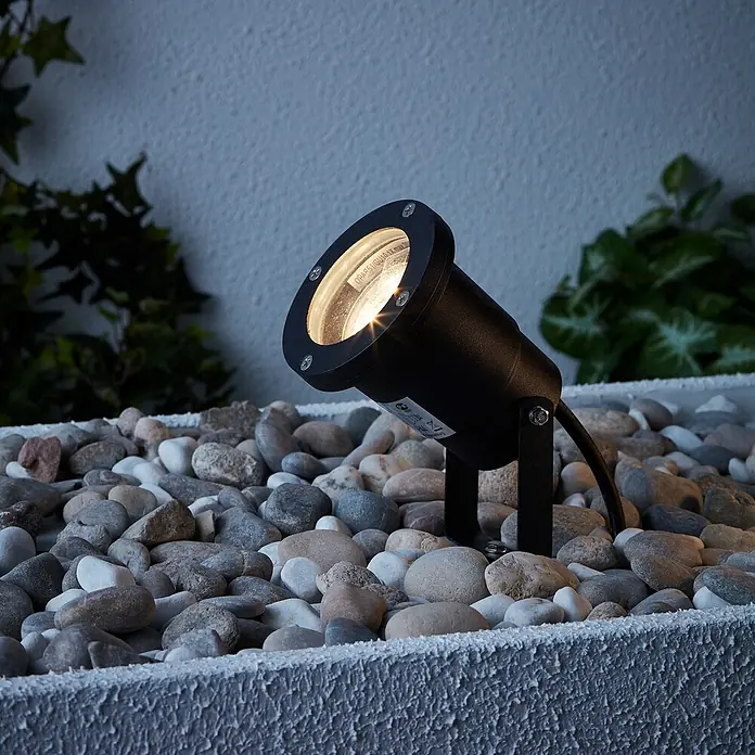 Brilliant LED-Erdspießleuchte Janko - 3 W, Ø x H: 9 x 16 cm, Schwarz, IP44 - Ein schwarzer, moderner Gartenstrahler steht auf Kies und beleuchtet dezent den Außenbereich.