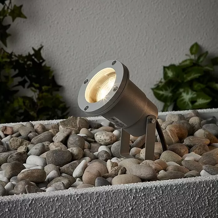 Brilliant LED-Erdspießleuchte Janko - 3 W, Ø x H: 9 x 16 cm, Grau, IP44 - Grauer Außenstrahler, warm leuchtend, eingebettet in Zierkies in einer Gartenumgebung.