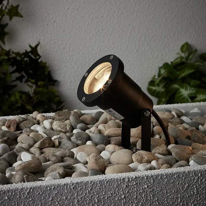 Brilliant LED-Erdspießleuchte Janko - 3 W, Ø x H: 9 x 16 cm, Schwarz, IP44 - Nahaufnahme einer schwarzen Gartenleuchte, die zwischen Steinen in einem Beet platziert ist und warmes Licht ausstrahlt.